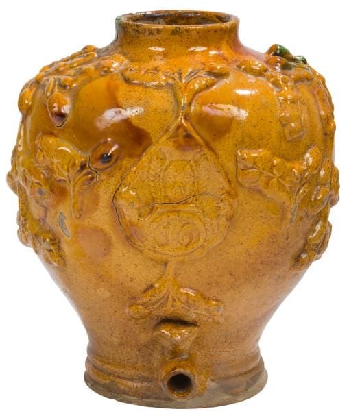 Occitanie, Toulouse, circa : 1814-1830 - 
Fontaine séditieuse en terre vernissée, réalisée par ou pour un soldat ayant servi dans le 16e Régiment d’Infanterie sous le 1er Empire et sous la Restauration (Régiment aussi dit « du Béarn » ayant participé à la bataille de Yorktown lors de la guerre d’Indépendance des États-Unis). Terre claire avec engobe, vernissée jaune, traces de manganèse, coulure partielle de vert. Décor de branches d’olivier autour d’une plaque de shako du 16e RI. Tête de Napoléon avec le bicorne, réalisée de façon séditieuse, au-dessus du robinet.
Hauteur : 30 cm