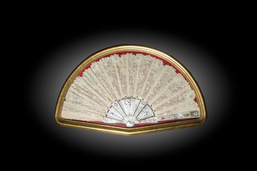Volutes, vers 1890-1800
Grand éventail, la feuille en dentelle à décor de volutes à modes variés et de fleurs. Huit grandes roses en bordure à triple pétale détaché. 
Monture en nacre blanche gravée de trois cartels dont au centre une femme en compagnie de deux amours. Sur les côtés, l’amour jardinier et sa bêche. Fine bordure dorée.
H.t. 32,3 cm / H.f. 19 cm (petites taches sur la feuille et petits acc. à la monture)
Dans un cadre à la forme, avec une baguette dorée (65,5 cm x 40 cm)
