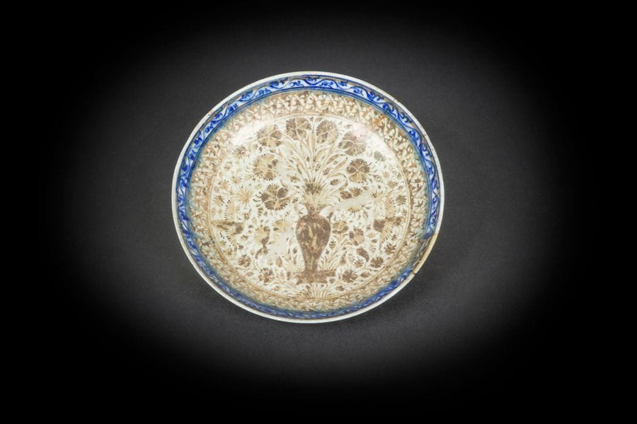 Plat au vase d’œillets, Iran safavide, fin XVIIe - déb. XVIIIe siècle
Céramique siliceuse sur petite base à décor à reflets métalliques et rehauts bleus sur fond crème. Vase entouré de tiges florales et oiseaux simurgh. Deux frises de motifs de palmettes et arabesques sur le rebord. Revers orné d’une frise de bouquet et fleurs épanouies alternées.
Anciennes étiquettes au dos : collection Mutiaux n°8 et 326 Perse (XVIIème siècle).
Diam. : 23,5 cm
État : un manque au rebord.

A Safavid pottery dish, Iran, late 17th/early 18th century