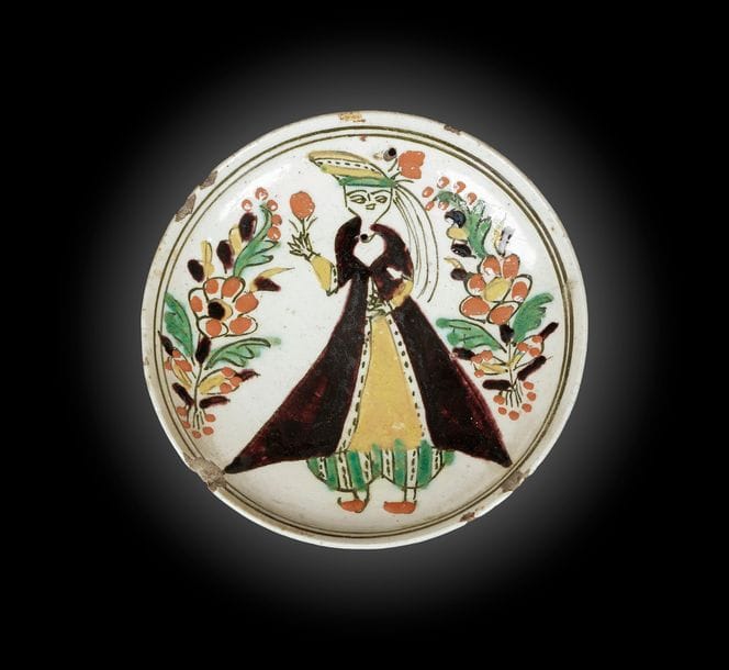 Coupelle à la jeune femme, Turquie ottomane, Kütahya, XVIIIe siècle
Céramique siliceuse peinte à main levée en polychromie sur engobe blanc et sous glaçure transparente. Une jeune femme, portant une tunique brune ceinturée sur un salwar à rayures vertes et pointillées noires, et un turban assorti, tient une fleur en main droite. Des motifs composites floraux l’encadrent et deux lignes noires au bord encerclent le tout.
D. : 15 cm ; H. : 4 cm
État : Bord ébréché, base fracturée et deux petits trous sur le fond.

A Kutahya pottery dish, Ottoman Turkey, 18th century