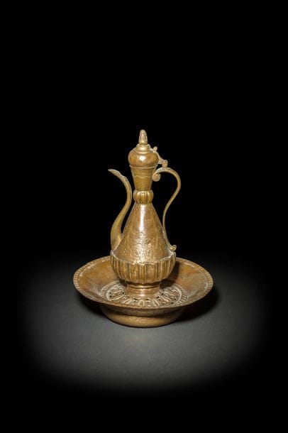 Aiguière, bassin et porte-savon en cuivre doré, tombak, Turquie ottomane, fin XVIIIe-déb. XIXesiècle
Aiguière à panse piriforme à col annelé à godrons et couvercle en dôme à prise en forme de pomme de pin. Décor ciselé de tiges florales tête-bêche sur un fleuron folié. Base goudronnée. Porte-savon, sabunlunk, à décor ajouré ceinturé d’une frise ciselée de compartiments. Le bassin reprend le même décor sur les bords.
H. aiguière : 33 cm ; D. bassin : 29,4 cm
État : dorure en partie effacée sur le bassin.

An Ottoman gilt-copper, tombak, ewer and matching basin, Turkey, late 18th- early 19th century.