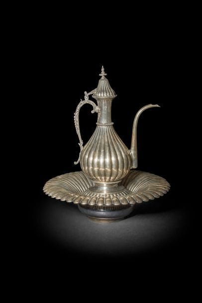 Aiguière et son bassin porte-savon, en argent, Empire ottoman, Égypte, fin du XIXe - déb. XXe
Très belle aiguière ibrik à panse piriforme munie d’une anse foliacée et d’un couvercle surmonté d’une prise en forme de fleuron. Panse à décor godronnée, ceinte d’un anneau perlé au col. Le porte-savon à godron est muni d’une grille à décor rayonnant ajouré et porte la couronne du khédive d’Egypte ‘Abbas Hilmï gravée à l’épaule. Le bassin évasé est également godronné. Porte une tughra difficilement lisible et le poinçon sah.
H. aiguière : 36 cm, D. bassin : 40 cm

Il est difficile de lire le nom de la tughra du sultan (Abdulhamid II (1876-1909)). Le khédive ‘Abbas Hilmi était vice-roi d’Egypte sous le sultan ottoman entre 1892 et 1914, date à laquelle il fut déposé par les Britanniques.

A silver ewer (ibrik) and basin, Egypt, late 19th/early 20th century