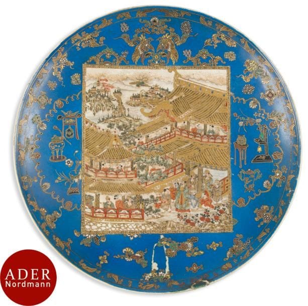 JAPON, Fours de Satsuma
Époque MEIJI (1868 - 1912)
Plat en faïence de Satsuma à décor émaillé polychrome et or à décor dans un panneau central d’une scène de palais avec enfants et personnages sous les pins, sur fond bleu à décor d’objets mobiliers et phénix. 
Diam. : 39 cm (éclats et restaurations).