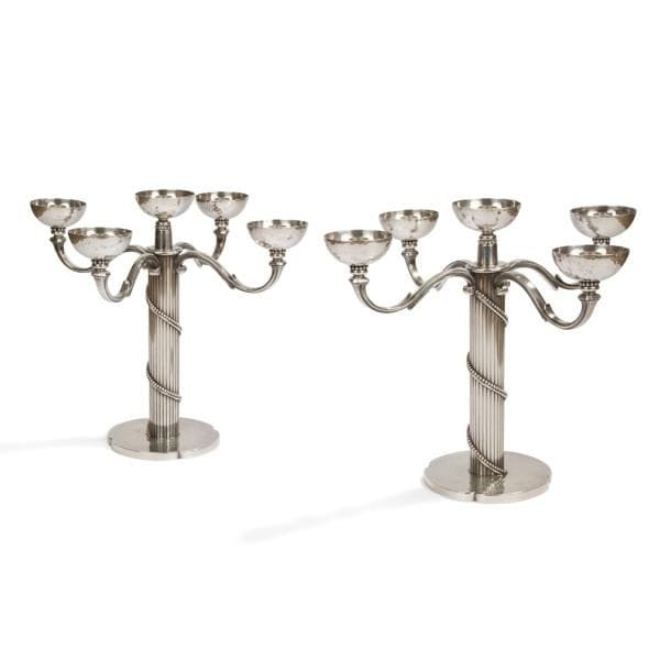 JEAN DESPRÉS (1889-1980)

Rare, élégante et importante paire de candélabres à cinq feux ; quatre bras de lumière et une bobèche centrale. 
Épreuves réalisées en métal argenté.
Les fûts cylindriques aux motifs d’épaisses cannelures sont chacun parcourus d’un rang perlé doublé d’un jonc en enlacement et reposent sur des bases martelées et quadrilobées.
Les bras de lumière en volutes et les fûts reçoivent des bobèches demi-sphériques martelées reposant sur de discrets rangs perlés.
Chaque candélabre signé J. DESPRÉS, à la pointe sous la base. 
Hauteur : 33,5 cm
Envergure : 40 cm

Provenance :
Ancienne Collection M. et Mme L. à Saint-Léger-Vauban, anciens commerçants avallonais. Œuvres acquises par ces derniers directement auprès de l’artiste.
