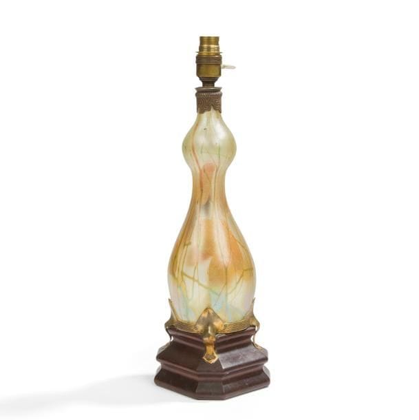 LOUIS COMFORT TIFFANY (1848-1933) ATTRIBUÉ À

Favrile Glass, série initiée en [1893]
Rare pied de lampe de forme bouteille ou vase monté anciennement.
Le corps, en verre richement irisé et au décor évoquant des feuilles et des tiges, repose sur un cerclage en laiton doré aux motifs de vaguelettes accueilli par une base en acajou sculpté.
Le col reçoit une bague en bronze patiné brun traitée dans le goût Nabi équipée du système électrique.
Circa 1900.
Sans accès à une possible signature masquée par la base couvrante en laiton doré.
Hauteur totale : 44 cm