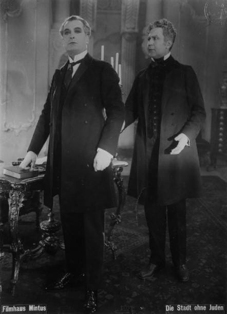 [PHOTOGRAPHIES - CINEMA] - DIE STADT OHNE JUDEN (la Ville sans Juifs) - Important ensemble de vingt-trois photographies de tournage du film « Die Stadt ohne Juden » (Filmhaus Mintus), d'après le roman de Maximilian Hugo Bettauer paru en 1922, certaines colorisées à l'époque, environ 23 x 30 cm chacune (petits trous de fixation aux angles, quelques doubles). La première de ce film expressionniste autrichien de Hans Karl Breslauer, tiré du livre qui est une véritable anticipation de ce qu'allait amener le nazisme (on entasse les juifs pour les expulser dans des fourgons à bestiaux, etc ...), eut lieu à Vienne le 25 juillet 1924 et l'auteur fut assassiné quelques jours plus tard par un nazi. Le livre a été traduit en français et publié en 1929 par Albin Michel. L'ensemble