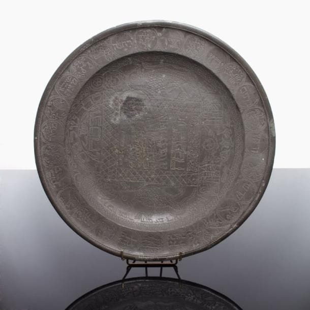 PLATEAU DE SEDER - Étain gravé au tremblé. Grand plateau au décor exubérant, gravé sur le marli d'une importante illustration animalière et humaine, avec les signes du zodiaque légendés en hébreu et les séquences du séder dans des médaillons, et au centre, entre deux lions extrêmement naïfs, une scène de Pessah et d'importantes inscriptions hébraïques relatives au « sacrifice de Pessah », avec une iconographie peu courante dans laquelle on découvre même un angelot à la partie inférieure et une date « 98 » sur le marli. France ou Allemagne, fin du XVIII ème siècle. Deux petits trous à l'étain, qui a été oxydé et restauré. Diam.: 43,5 cm.