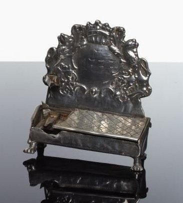 LAMPE DE HANOUCCA - Étain moulé et gravé. La lame porte-godets, recouverte d'un couvercle à croisillons (manque une partie de ce couvercle ; godets probablement remplacés) est placée dans un corps en étain qui repose sur quatre pieds griffes. Poinçons illisibles (alsaciens ou allemands), sous le corps en étain. Le dosseret à décor rocaille floral est orné de deux lions supportant une couronne ouverte. Inscription au centre du dosseret M 1737. Beau travail du XVIII ème siècle, probablement de la vallée du Rhin. Haut.: 15 cm. Long.: 19 cm.