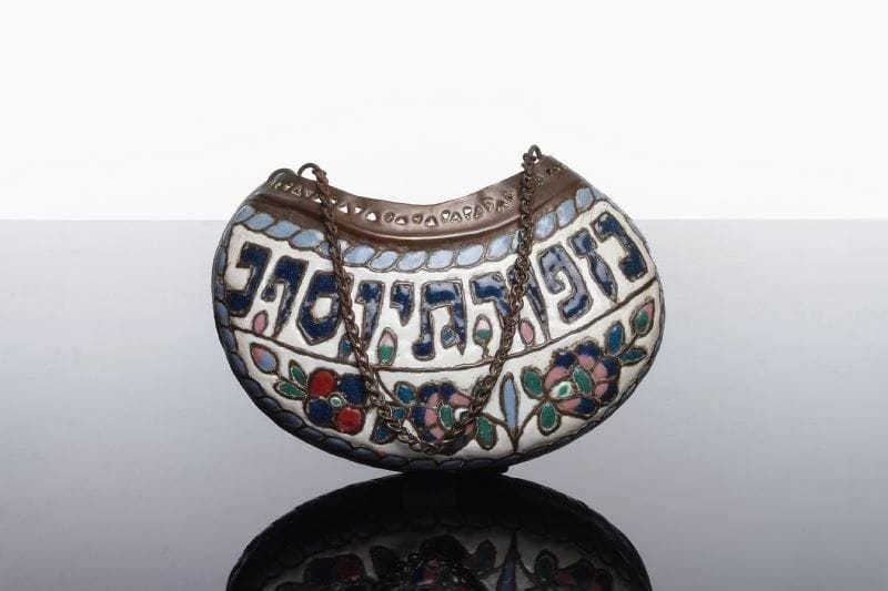 SUSPENSION - Cuivre ajouré et émaillé, chaîne en métal. Le décor polychrome comporte sur chaque face d'un corps en forme de haricot, au-dessous d'une inscription hébraïque porte-bonheur (Joseph ben Porat ...), une frise de fleurs et feuillages. Afrique du Nord ou Moyen Orient, XIX ème-XX ème siècle. Long.: 19 cm. Haut (sans la chaîne): 11 cm (petites écaillures à l'émail).