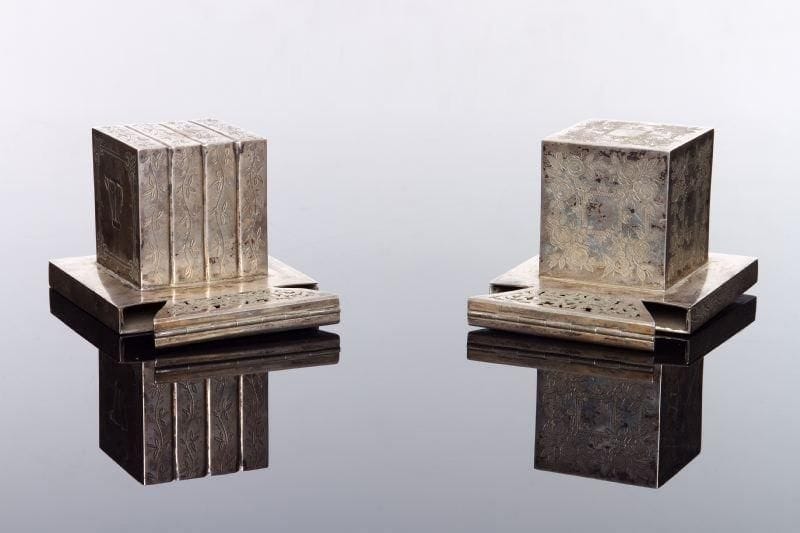 PAIRE D'ÉTUIS À TEFILIN - Argent ajouré et gravé d'un important décor floral sur toutes les faces, l'un des étuis avec la lettre Shin répétée en relief sur deux faces. Bien que les décors floraux latéraux soient différents sur les deux étuis, l'identité des décors à la partie inférieure et sur la partie ajourée laisse penser qu'il s'agit d'une vraie paire. Ancien poinçon viennois peu lisible (1859 ?) sur les étuis Haut.: 7 cm. Long.: 8 cm. Poids total: 421 g.