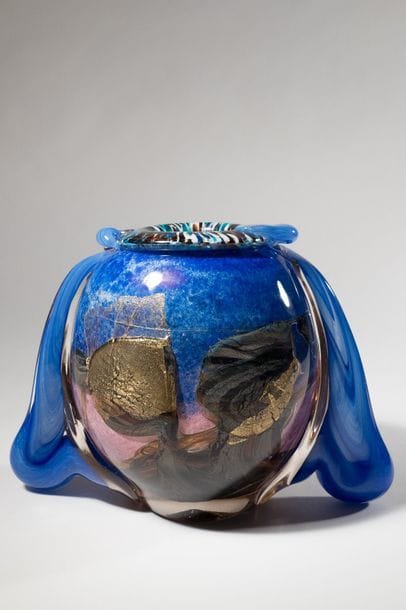 Jean-Claude NOVARO (1943-2015)
 Vase boule en verre soufflé bleu et rose, à décor de cordons internes marron et feuilles d’or, deux larges anses de verre bleu l’enserrant.


Signé et daté 2007 sous la base.


Hauteur : 22 cm.