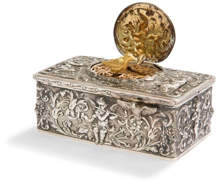 Boîte à musique en argent (800 ‰) à oiseau chanteur en vermeil. De forme rectangulaire elle reçoit sur toutes les faces un décor de putti ailés jouant pour la majorité de différents instruments à vent dans des enroulements feuillagés et fleuris. Dans la cuvette du revers le décor s’articule autour d’un vase Médicis rempli de fleurs encadré de deux paons. Le bouton poussoir figure un oiseau les ailes déployées.
ALLEMAGNE fin du XIXe siècle, début du XXe siècle 
État : à restaurer, manque la clé.
Poids brut : 461,70 g - longueur : 10 cm - largeur : 5,7 cm - hauteur : 3,8 cm