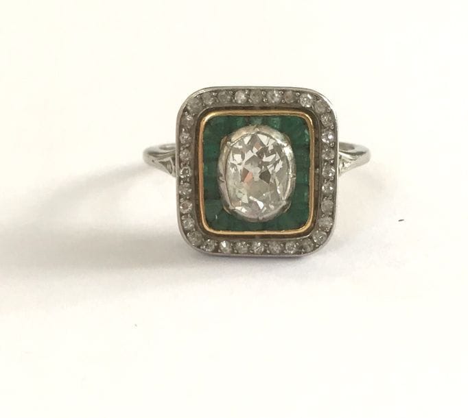 Bague en platine et en or gris 18K (750), ornée d’un diamant de forme ovale entouré d’émeraudes calibrées ceintes de diamants ronds de taille ancienne. Travail français des années 1925. Tour de doigt : 54. 
Poids brut : 4,8 g