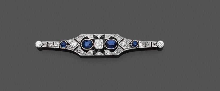 Broche fuseau en or gris 18K (750) et platine, ajourée de motifs géométriques imbriqués, centrée d’un diamant de forme coussin entre deux saphirs ovales, la monture sertie de diamants ronds, agrémentée de deux saphirs ronds. Travail français des années 1925. Longueur : 7,5 cm environ. 
Poids brut : 8,9 g