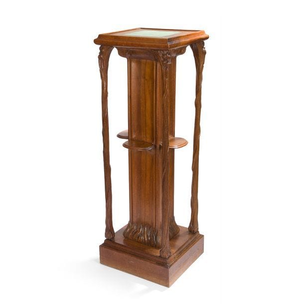 TRAVAIL D’ÉPOQUE ART NOUVEAU
Étonnante sellette naturaliste en noyer mouluré et sculpté.
Le fût central, de section carrée, est agrémenté en son centre, sur trois de ses faces, d’une tablette et repose sur une épaisse base à un léger gradin.
Il est souligné d’une moulure à chaque angle simulant une tige florale ; sa partie basse et son chapiteau sont aux motifs sculptés de feuilles lancéolées.
Le plateau, à la bordure moulurée, accueille, en chute jusqu’à la base, des montants arborescents sculptés enrichis de motifs floraux en partie haute.
Le dessus est constitué de neuf carreaux en céramique émaillée vert.
Circa 1900.
119 x 40 x 40 cm