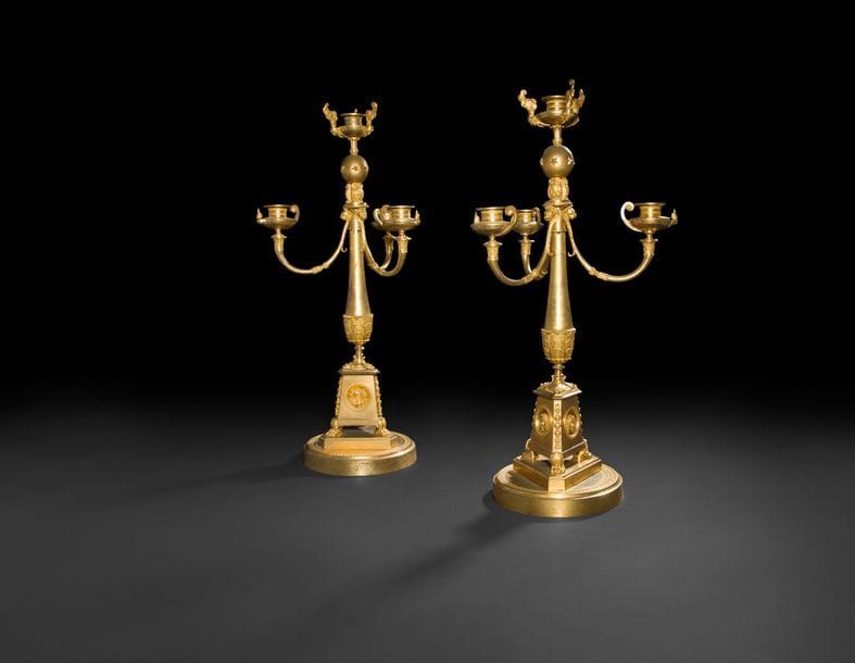 Rare paire de candélabres en bronze doré à quatre lumières à bobèches à lampes antiques et nuées à mascaron reposant sur une sphère étoilée supportée par trois hiboux surmontant des mufles de lion au sommet d’un fût en balustre à base triangulaire ornée de masques de Diane, têtes de chimère et pieds en griffe, le socle circulaire à décor guilloché.
Attribués à Claude Galle ou Louis-Auguste Hervieu
Époque Consulat (usures).
H?: 56,5?cm 

 Le motif du hibou se retrouve sur une paire de flambeaux livrés par Claude Galle en 1804 au château de Fontainebleau (J-P. Samoyault, Musée national du château de Fontainebleau, Pendules et bronze d’ameublement entrés sous le Premier Empire, Paris, 1989, p.176, n°157). Par ailleurs une mention citée dans un catalogue de vente (Vente Neuilly, 11 juin 2012, lot 169) fait état d’une paire de flambeaux à hiboux en 1799 dans l’atelier de Claude Galle. D’une manière plus générale, ces candélabres évoquent un autre modèle souvent réalisé en bronze patiné et doré présentant également une base triangulaire et trois hiboux surmontant une sphère, attribués par Jean-Dominique Augarde au bronze Louis-Auguste Hervieu (1765-1811) et dont un exemplaire a récemment été vendu à Paris, étude Brissonneau, le 21 mars 2018, lot 259 (Voir J.D. Augarde, Une nouvelle vision du bronze et des bronzier sous le Directoire et l’Empire, L’estampille l’objet d’art, n°398, p. 80-84).