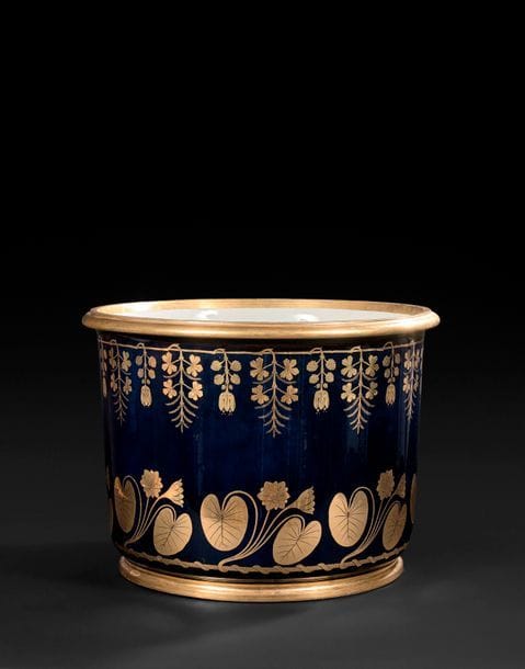SèVRES
Grand seau à laver les pieds cylindrique en porcelaine dure à décor sur fond bleu en or de guirlandes de nymphéas alba et guirlandes de renoncules aquatiques et feuillages.
Marqué Manufacture impériale de Sèvres en rouge pour 
An XIII (novembre 1804 - novembre 1805)
H : 35,5 cm, D : 49,5 cm
(Petits éclats à la base). 

Provenance : 
- Livré en décembre 1806 par ordre de l’Empereur Napoléon Ier en présent au Grand-Duc Charles II de Bade par l’intermédiaire de Monsieur le baron de Gensau, conseiller de légation à la cour de Bade à l’occasion du mariage de la nièce adoptive de l’Empereur, Stéphanie de Beauharnais avec le Grand-Duc Charles II de Bade. 
- Sammlung der MarkgraFen und Grossherzöge von Baden, Sotheby’s, 16-30 octobre 1997, lot 4919. 

Ce seau entre au magasin de vente de la manufacture de Sèvres le 9 floréal an XIII (30 avril 1805) au prix de vente de 1 200 francs ainsi décrit : Un seau à laver les pieds, frise du bas Nymphea alba, bord supérieur, renoncule d’eau aquatique. Deux dessins anonymes pour des frises sont conservés aux archives de la manufacture de Sèvres.
Un autre seau à laver les pieds, de la même forme est peint en 1784 d’une frise de rinceaux feuillagés or sur fond beau bleu. Ce seau daté 1784 est resté en stock à la manufacture au XVIIIe siècle puis il est offert en 1801 par Bonaparte au roi d’étrurie (information aimablement communiquée par John Whitehead). Puis il est mentionné dans les collections du Roi George IV d’Angleterre à Carlton house en 1826 et aujourd’hui au musée du Louvre (Christie’s, Londres, 6 octobre 1986, lot 256 et Pierre Ennès, Nouvelles acquisitions du département des Objets d’art, 1990-1994, musée du Louvre, pp. 204-205, n°86).
Notre seau est livré en décembre 1806 par ordre de l’Empereur à monsieur le baron de Gensau, conseiller de légation à la cour de Bade à l’occasion du mariage de la princesse Stéphanie de Beauharnais avec le Grand-Duc Charles II de Bade puis resté dan