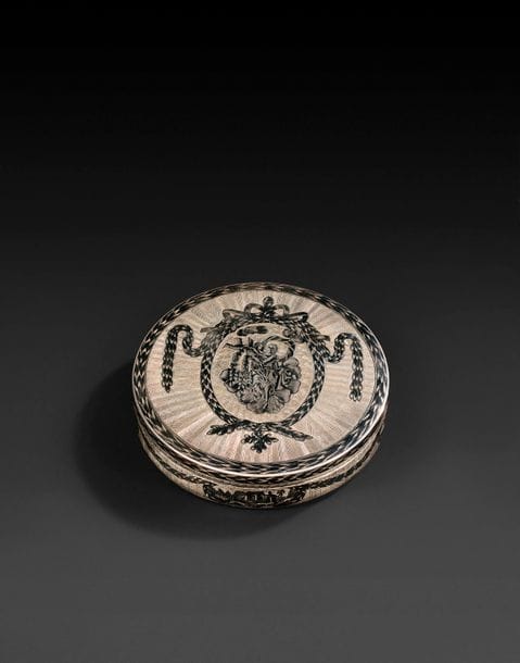 Tabatière ronde légèrement bombée, en argent et vermeil à décors niellés.
Le couvercle représente, au milieu de nuées, Aphrodite tendant les bras à Éros. Ce décor est gravé sur un fond guilloché, dans un médaillon ovale souligné de guirlandes au milieu de rayons dorés. Le bord est souligné d’une couronne tressée de feuillage. Le fond présente la même structure, avec au centre Éros tenant dans chaque main un rameau de palme. La ceinture est aussi richement décorée de quatre médaillons ovales soulignés et reliés entre eux par une guirlande de feuillage. L’intérieur est en vermeil.
Poinçons : 84, Moscou, 1827, essayeur « НД » (Nikolaï Loukitch Doubrovin, (? -1862), actif en 1822-1855) et orfèvre « БГ » (non identifié)
Diamètre : 9,3 cm ; Hauteur au centre : 2,3 cm
Poids brut : 160,81 g

Une tabatière rectangulaire en argent et vermeil à décors niellés avec des poinçons identiques a été vendue chez Gelos en 2011.
 
Серебряная круглая табакерка с чернью, слегка выпуклая сверху и снизу.
На крышке изображена Афродита сидящая на облаке, принимающая в объятия летящего Эроса. Гильошированный фон, овальный медальон в растительной рамке. Сама рамка на фоне гравированных позолоченных лучей. Край крышки подчёркнут гирляндой из листьев. На дне аналогичный декор и изображение Эроса, сидящего на облаке с пальмовой ветвью в каждой руке.
Пояс табакерки также богато украшен четырьмя овальными медальонами в обрамлении из листовой гирлянды и соединены между собой подобной гирляндой.
Внутри позолочена.
Клейма : Москва, 1827 г., пробирный мастер «НД» (Дубровин Николай Лукич, умер в 1862 г., работал в 1822 - 1855 гг.) и мастер «БГ»
Диаметр : 9,3 см ; Высота посередине : 2,3 см ; Общий вес : 160,81 г.

Серебряная прямоугольная табакерка с позолотой и с чернью с такими же клеймами была продана у Гелоса в 2011 г.

 
