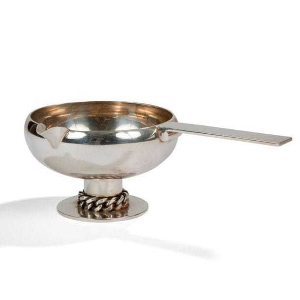 JEAN DESPRÉS (1889-1980)
Élégante saucière en métal argenté. Base circulaire martelée et ornée d’une gourmette à maillons plats. Prise ornée d’un cylindre et à surface martelée. Signée J. DESPRÉS à la pointe et porte le poinçon de Maître Orfèvre au revers de la prise. 7 x 20,5 x 13 cm
