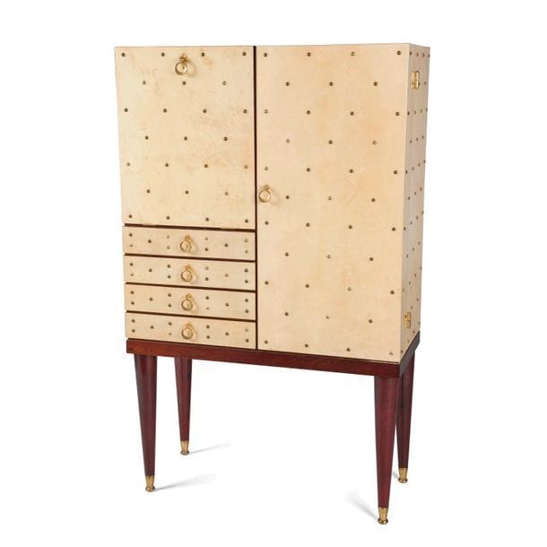 ATTRIBUÉ À L’ATELIER ALDO TURA
Cabinet formant meuble-bar en parchemin verni au décor de cloutage en laiton et à l’intérieur éclairant. La façade s’ouvre, à droite par un caisson découvrant un intérieur muni de tablettes en verre sur fond de miroir. La partie gauche présente un abattant, découvrant également un intérieur fait de miroir et de verre, surmontant une série de quatre tiroirs. Base et pieds coniques en acajou. Sabots de pied et prises en métal doré. Années 1960. État d’usage, petits éclats de vernis épars. 130 x 80 x 36 cm 