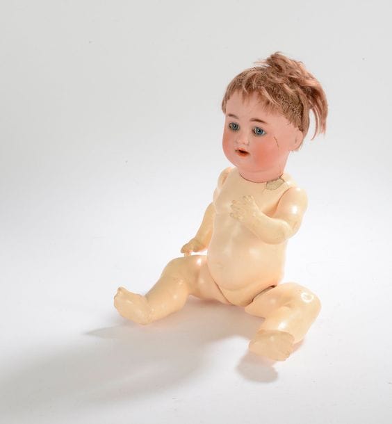 Petite poupée allemande, tête en porcelaine (accidentée et recollée), yeux fixes bleus, bouche ouverte sur 2 dents, perruque blonde, corps raide en composition.

Hauteur : 35 cm