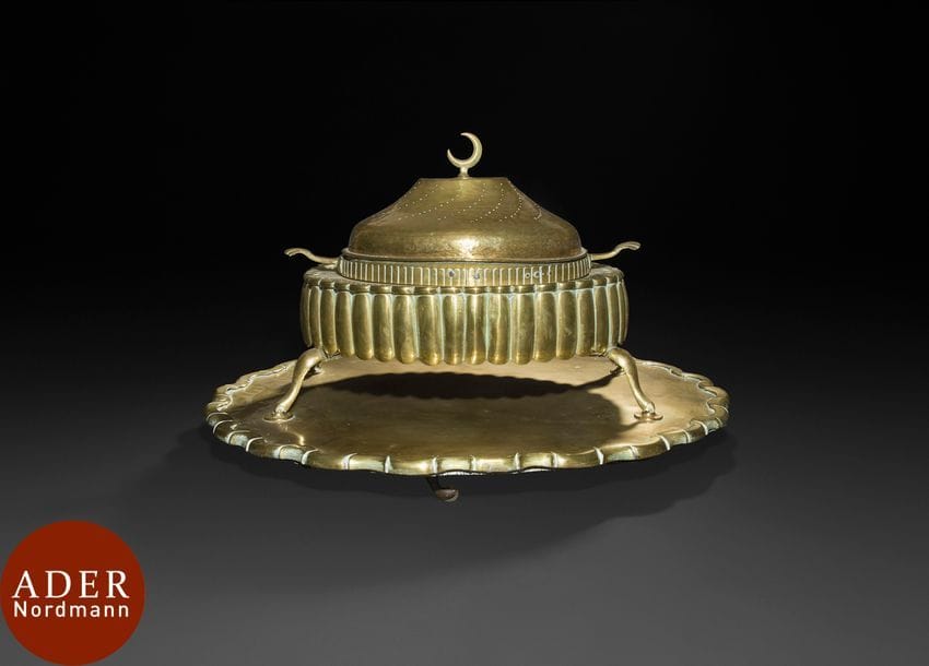 Brasero ottoman en laiton, Turquie, fin XIXe 
Brasero à base godronnée reposant sur quatre pieds, cuve à braises amovible flanquée de deux poignées coquille et couvercle convexe à décor de courbes ajourées et d’un croissant de lune au sommet. Accompagné de son plateau à roulette en laiton doré à découpe festonnée. 
Dim. : 63,5 x 45 x 47 cm
État : quelques rayures, coups, sauts de dorure.

An Ottoman brass brasero, late 19th century