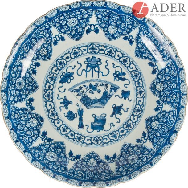 CHINE - Époque KANGXI 
(1662 - 1722)
Plat rond en porcelaine blanche polylobé à décor central émaillé en bleu sous couverte dans un médaillon d’objets mobiliers et d’une femme et bouquet fleuri dans un éventail, de fleurs de lotus dans leur feuillage en réserve. (Éclat se transformant en fêlure, petites égrenures en bordure).
Diam. : 38,4 cm
