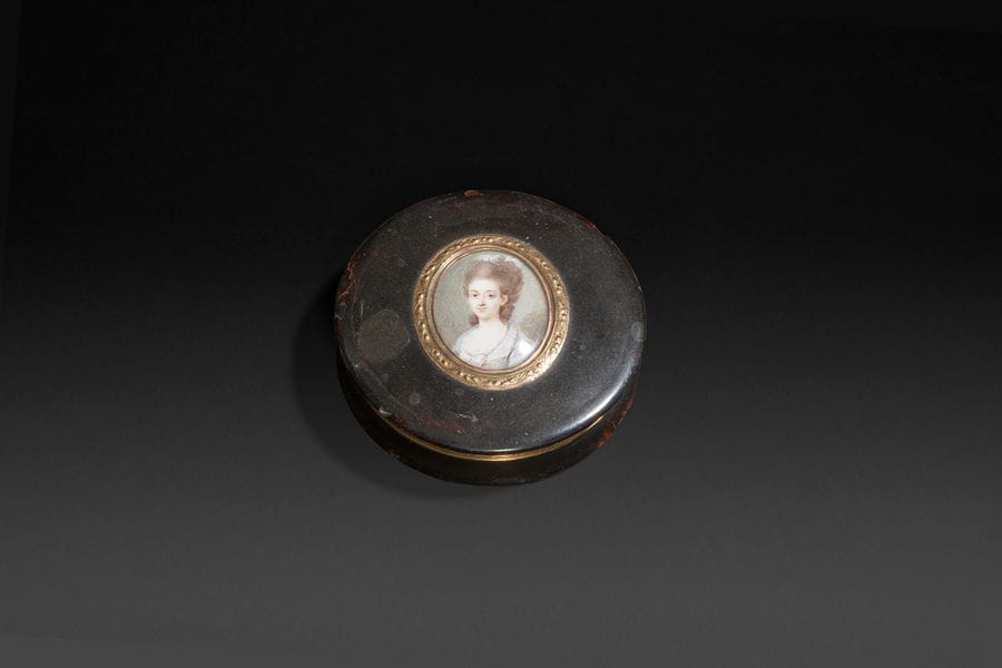 École française du XVIIIe siècle
Portrait de jeune femme, en buste de trois-quarts à gauche, des plumes d’autruche dans sa haute coiffure, en robe bleu pâle.
Miniature sur ivoire vers 1775, à vue ovale 4,5 x 3,7 cm, entourée d’or de deux couleurs à motif de nœuds de ruban sur le couvercle d’une boîte du début du XIX siècle en écaille brune montée en or et à la bâte incurvée, diam. 8,3 cm, P. 2 cm.
Poinçons sur la monture, dont celui du maître orfèvre parisien Jean-Ange-Joseph Loque, insculpé en 1798 (initiales JL et une balance, dans un losange vertical).
et poinçon au coq.
Poids brut : 51,40 gr.
(nombreux petits accrocs à la boîte).