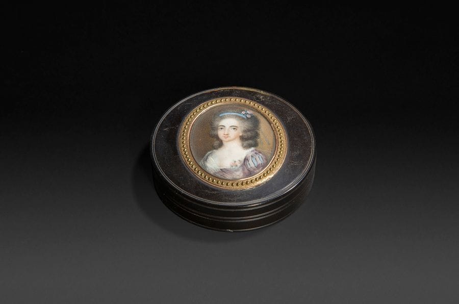 École française du XVIIIe siècle
Portrait de jeune femme, un ruban bleu et trois roses dans sa coiffure bouffante, en robe violine, manches à soufflets, des roses au corsage, à mi-corps vers la gauche sur fond brun.
Miniature sur ivoire vers 1785, à vue ovale 4,5 x 3,7 cm, entourée d’or de deux couleurs à motif perlé sur le couvercle d’une boîte en écaille du XVIIIe siècle, diam. 7,2 cm, P. 1,8 cm.
(verre irisé ; légères abrasions au couvercle)