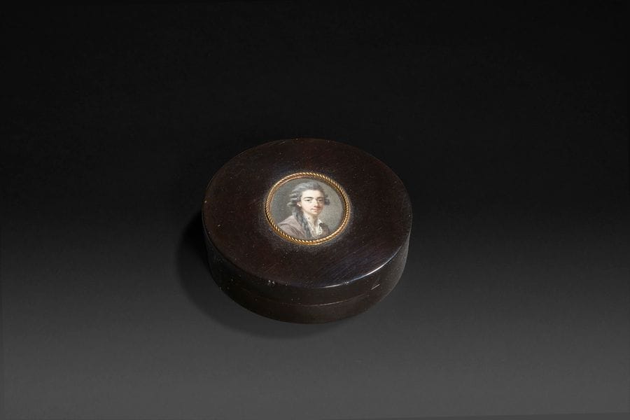 Nicolas LAFRENSEN dit LAVREINCE (Stockholm, 1737 - 1807).
Portrait d’un jeune homme à la longue natte, en veste marron à grand col sur chemise à col ouvert, un artiste ?
Miniature sur ivoire du XVIIIe siècle, signée indistinctement en bas à droite : Laf 
ovale à vue 3 x 2,6 cm, cerclée d’un filet torsadé en or sur le couvercle d’une boîte du XVIIIe siècle en écaille, diam. 7,4 cm, P. 1,9 cm.
(Examinée sans démontage; poussières sous le verre, très petites abrasions à la boîte) 

Peintre en miniatures du roi de Suède et de l’Académie de Stockholm, qui fit une grande partie de sa carrière en France où il fut fameux pour les sujets en miniature et à la gouache.