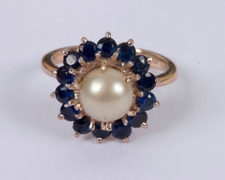Bague en or gris 18K (750), ornée d'une perle bouton fantaisie entourée de pierres bleues. Tour de doigt : 48. Poids brut : 5,2 g