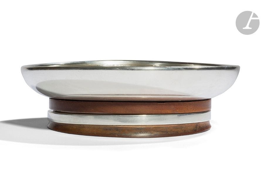 JEAN-ÉMILE PUIFORCAT (1897-1945) (JEAN-ÉLISÉE PUIFORCAT, DIT)
Coupe sur talon en argent et palissandre.
Le talon composé de trois strates, deux en palissandre et une en argent, reçoit la coupe creuse achevée ourlée vers l’intérieur.
Déformations.
Signée Jean E. PUIFORCAT et porte les poinçons de Maître Orfèvre et de garantie sur la base.
Hauteur : 6,3 cm - Diamètre : 21,3 cm
Poids brut : 1.073 g