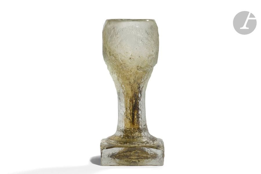 MAURICE MARINOT (1882-1960)
Sans titre, circa 1923
Vase calice à longue jambe reposant sur une épaisse base carrée.
Épreuve en verre miel très épais présentant une coloration interne bistre vers le centre ; la surface extérieure très profondément travaillée à l’acide sur sa surface, des parties éparses laissées lisses dont le col achevé à froid à la meule.
Signé MARINOT sous la base.
Hauteur : 18,7 cm

Bibliographie : 
Maurice Marinot artisan verrier - Catalogue raisonné de l’œuvre de verre - Félix Marcilhac, les Éditions de l’amateur, Paris, 2013. Notre vase pourrait être le vase, non illustré et non identifié, référencé sous le n° 700 (1923) à la page 365 et décrit comme suit : Cat. MM n° 700 (1923) – Œuvre non identifiée – Descriptif : Verre pied carré, givré partout. Dimensions non relevées. 

Provenance :
Ancienne Collection de Bormans. Collection non localisée. 

Œuvres en rapport :
Maurice Marinot artisan verrier - Catalogue raisonné de l’œuvre de verre - Félix Marcilhac, les Éditions de l’amateur, Paris, 2013.
- Grand verre à grande jambe de 1924 référencé sous le numéro 782 et décrit et reproduit page 380.
- Coupe à pied de 1932 sous le numéro 2176 et décrite et reproduite page 639.