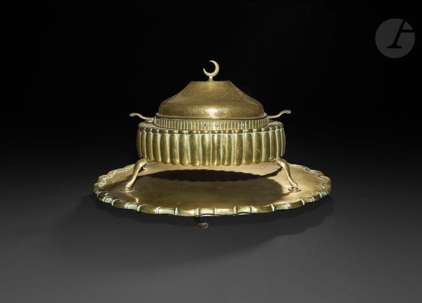 Brasero ottoman en laiton, Turquie, fin XIXe siècle
Brasero à base godronnée reposant sur quatre pieds, cuve à braises amovible flanquée de deux poignées coquille et couvercle convexe à décor de courbes ajourées et d’un croissant de lune au sommet. Accompagné de son plateau à roulette en laiton doré à découpe festonnée.
63,5 x 45 x 47 cm
État : quelques rayures, coups, sauts de dorure.

An Ottoman brass brasero, Turkey, late 19th century