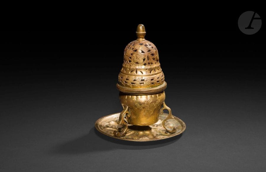 Brûle-parfum en tombak, Turquie ottomane, XVIIIe - XIXe siècle
Brûle-parfum en cuivre doré, tombak, en forme de dôme reposant sur une cassolette tripode rivetée sur un petit plateau circulaire plus tardif. Couvercle articulé à bouton de préhension en forme de gland et orné d’un décor floral ajouré et gravé de rosettes et feuillages. Plateau à décor repoussé de pétales tournoyants.
Hauteur : 19 cm
État : traces d’oxydation et d’usures de la dorure.

An Ottoman tombak incense burner, Turkey, 18th - 19th century