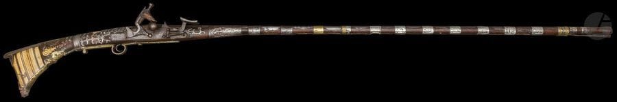 Fusil mukhala à platine à silex, Maroc, fin du XIXe siècle
Long canon en bois à bagues d’argent ciselées d’un décor de rinceaux, garnitures et deux bagues en laiton. Crosse et fût à garnitures d’os gravées, de plaques d’argent ciselé découpées en arabesques et de clous. Rehauts de peinture rouge et détente et pontet en laiton.
Longueur : 159,5 cm
État : accidents et petits manques.

A flintlock gun, mukhala, Morocco, late 19th century 