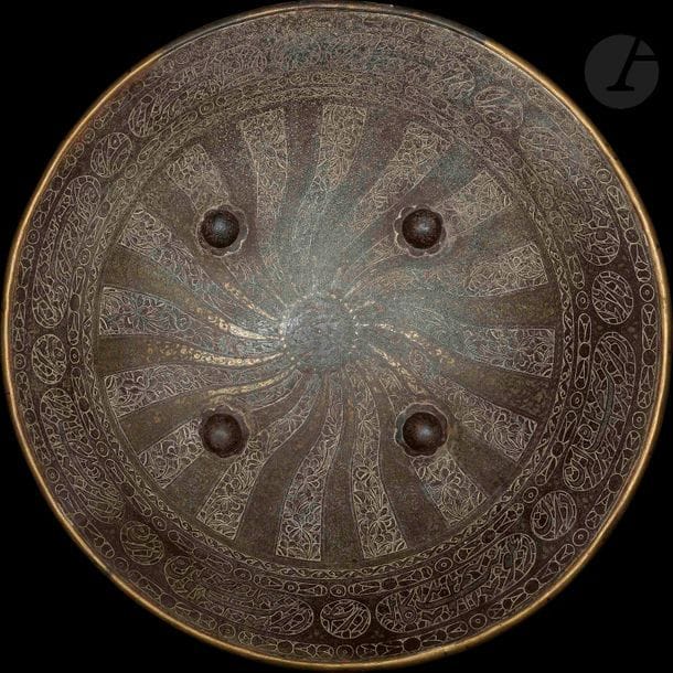 Rondache à quatre bossettes en alliage cuivreux, Iran qâjâr, XIXe siècle
Décor incisé et partiellement incrusté d’or de bandeaux floraux tournoyants autour d’un rondeau inscrit du nom de Shah Abbas. En bordure, frises de cartouches et rondeaux calligraphiés de vers de poésie persane.
Diamètre : 52 cm

A copper round shield, Iran, 19th century