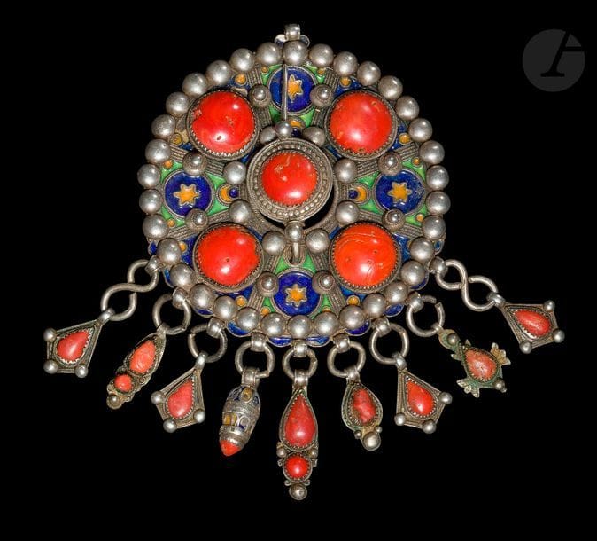 Grande fibule ronde, tabzmit, Algérie, Grande Kabylie, Beni Yenni, début XXe siècle
Grande fibule en argent ciselé, décorée d’émaux cloisonnés jaune, vert et bleu, sertie de quatre cabochons de corail et d’un autre au centre sur l’épingle de la fibule. Le dos orné d’une rosace stylisée. L’ensemble agrémenté de neuf pampilles serties de corail.
Diamètre : 12 cm ; Poids brut : 493,3 g

A large round enamelled silver fibulae, tabzmit, Algeria, Great Kabylia, Beni Yenni, early 20th century
