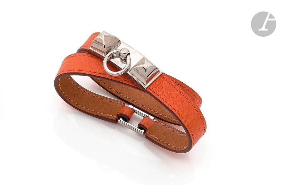 HERMÈS. Bracelet en cuir orange et motif Médor en métal. Signé. Dans sa pochette et sa boîte. Longueur : 36 cm environ