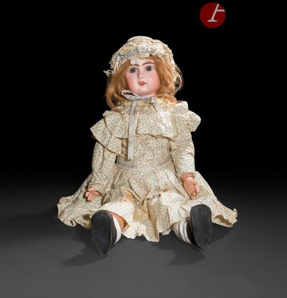 Grande poupée, tête en porcelaine marquée en creux : 13 (Jumeau tardive). Yeux fixes bleus, bouche ouverte sur une rangée de dents, oreilles rapportées et percées, perruque blonde. Corps articulé en bois et composition (petits accidents). Robe et bonnet en coton imprimé de fleurettes roses et bleues (taches).
H : 79 cm