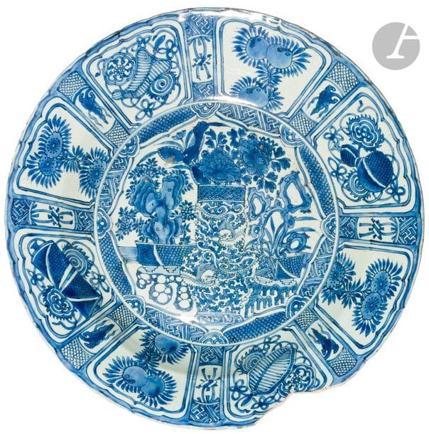 CHINE - Début XVIIe siècle
Grand plat Kraak en porcelaine émaillée en bleu sous couverte à décor de vases fleuris ornés de dragons et de rocher en décor central, de panneaux en réserve ornés d’objets bouddhiques, de fleurs, de pêches et de svastika. (Fêlures, éclats, sauts d’émail, égrenures). 
Diam. 50,5 cm