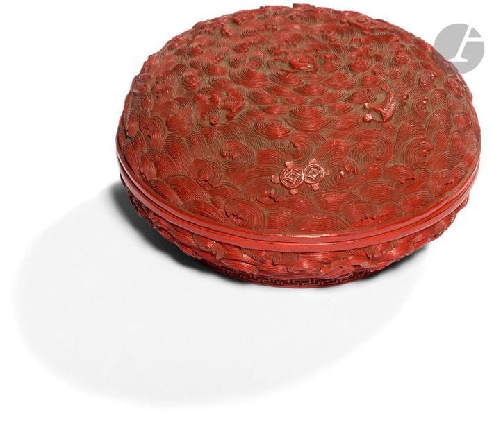 CHINE - Époque JIAQING (1796 - 1820)
Boîte ronde en laque cinabre rouge à décor ciselé en relief de vagues écumantes mouvementées surmontées d’un décor central de la perle enflammée et parsemé d’emblèmes bouddhiques. (Petites égrenures, gerces et usure de laque). 
Diam. 11 cm