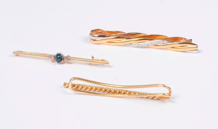 CARTIER. Pince-cravate en trois brins d'or 18K (750) de couleurs, celui en or gris agrémenté de diamants ronds. Signé et numéroté. Longueur : 6 cm environ. On joint 2 bijoux en or 18K : un pince-cravate et une broche-barrette agrémentée d'un saphir cabochon entre deux diamants taillés en roses. Poids brut : 19,6 g