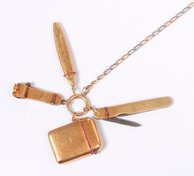 Longue chaîne en or 18K (750) munie de ses accessoires en or 18K aux armes de marquis : un étui à allumettes-pyrogène, un canif, un porte-mine et un coupe-cigare. Poids brut : 118,3 g (enfoncements, déformations)