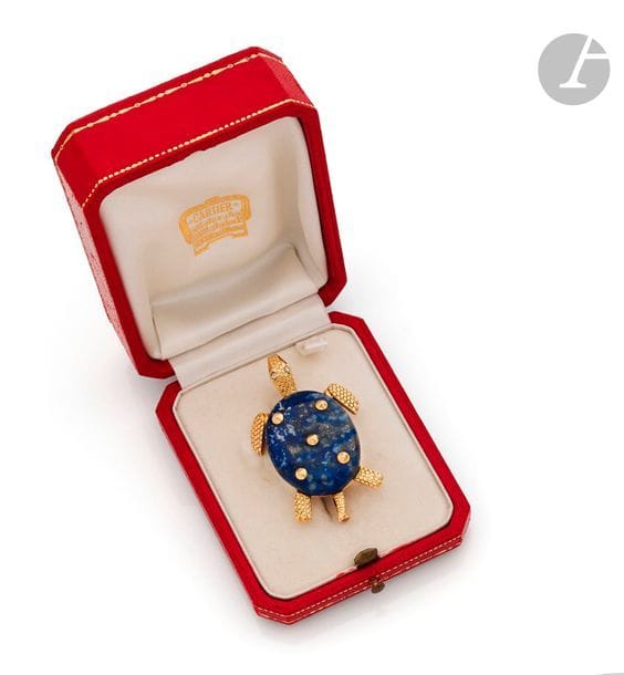 CARTIER
Broche en or 18K (750) dessinant une tortue, le corps en lapis-lazuli, les yeux soulignés d’un diamant taillé en rose. Signée. Travail français. Dans son écrin. 
Hauteur : 5 cm environ. 
Poids brut : 22,7 g