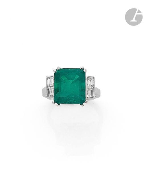 Bague en or gris 18K (750), ornée d’une émeraude pesant 6,08 cts de forme rectangulaire à pans coupés, épaulée de diamants calibrés. 
Tour de doigt : 53/54
Poids brut : 5,6 g
 L’émeraude accompagnée d’un rapport LFG (2019) attestant selon son opinion : origine Colombie, présence d’huile et de résine (choc)