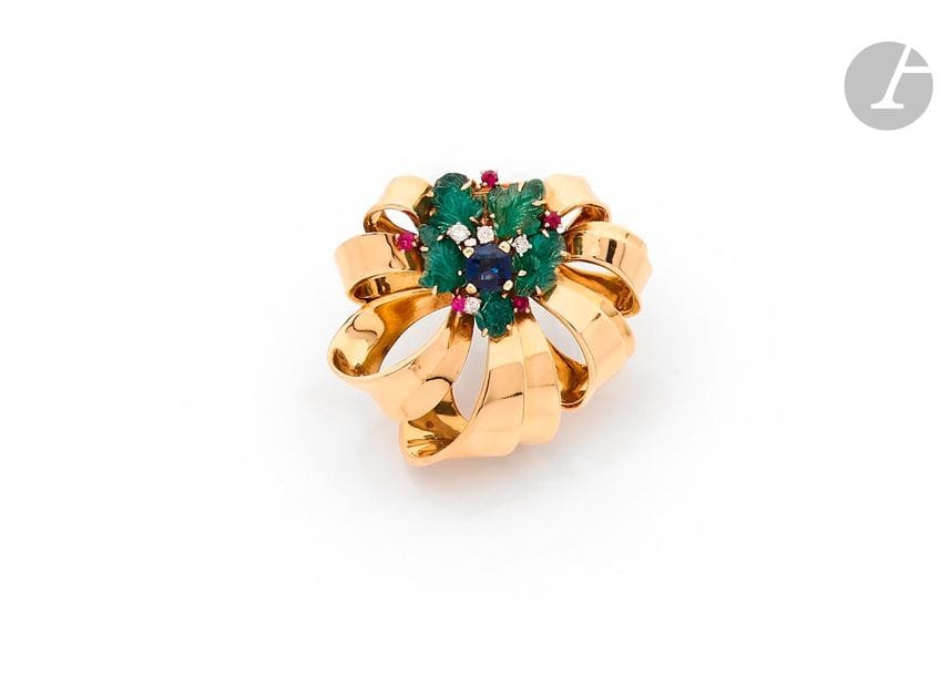 CARTIER
Clip en or jaune 18K (750) dessinant un nœud de rubans, orné d’un bouquet de feuilles en émeraudes gravées entourant un saphir de forme coussin, agrémenté de diamants ronds et de rubis. Signé. Travail français des années 1940. 
Dimensions : 4 x 4 cm environ. 
Poids brut : 21,3 g